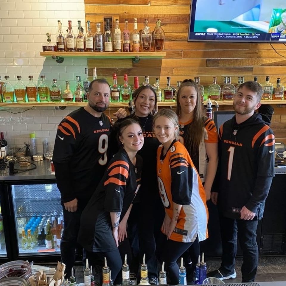 Five people in Cincinnati Bengals jerseys pose behind a restaurant bar lined with liquor bottles and televisions.