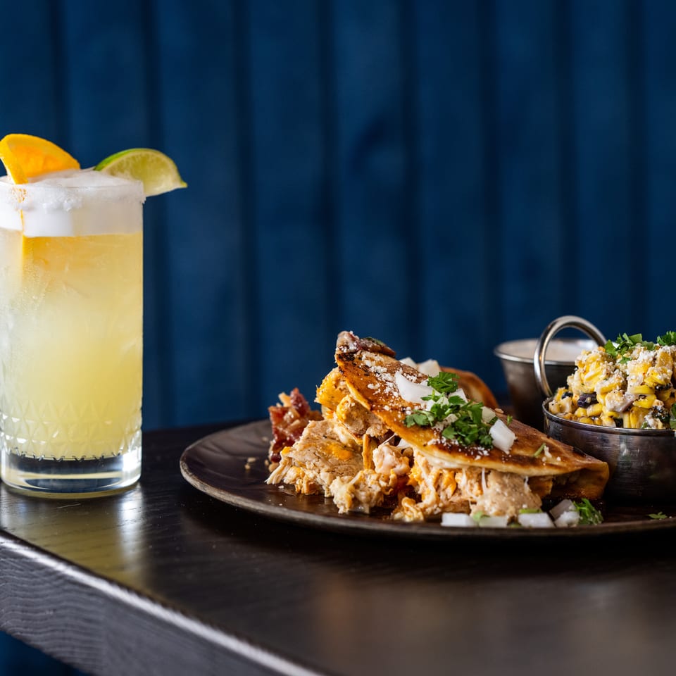 Tacos, corn salad, and a citrus cocktail with orange and lime garnishes arranged on a dark restaurant table.