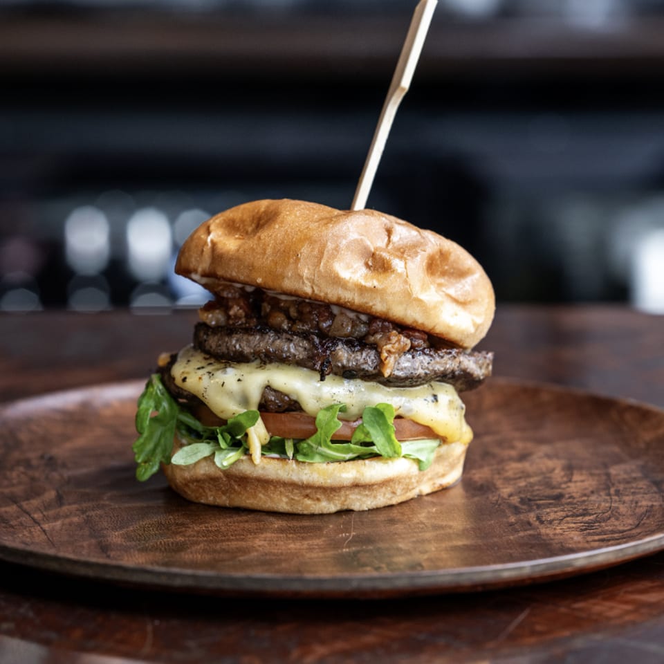 Gourmet burger with melted cheese, caramelized onions, arugula, and tomato on a wooden plate.