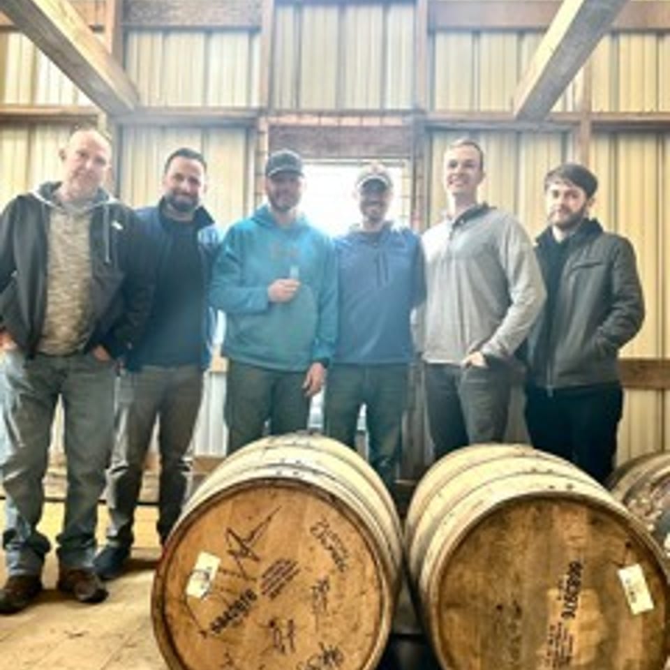 Six men stand behind two large wooden barrels inside a rustic barn with exposed beams.
