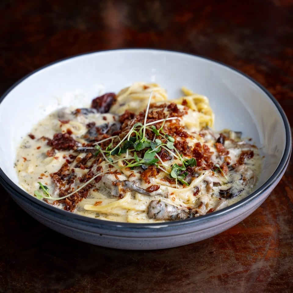 Creamy pasta with mushrooms, sun-dried tomatoes, bacon bits, and microgreens in a white bowl.