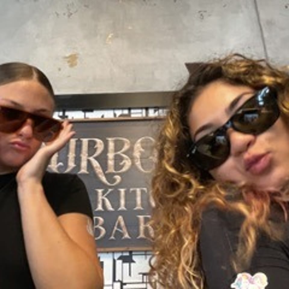 Two women wearing sunglasses pose in front of a restaurant sign, one with straight hair and one with curly hair.
